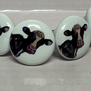 May include: Set of four ceramic cabinet knobs. Each knob features a white background with a black and white cow face design. The cow has a pink nose and a light brown patch of fur.