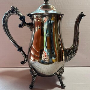 May include: A silver-colored, antique coffee pot with a lid and ornate handle. The pot has a curved spout and sits on three decorative feet. The reflective surface mirrors the surroundings, adding depth to the design. A classic piece for serving beverages.
