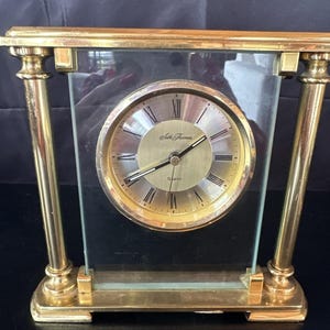 May include: A gold-toned quartz clock with a rectangular glass case. The clock face is gold with black Roman numerals and hands. The brand name "Seth Thomas" is printed on the clock face. The clock has a classic, vintage design.