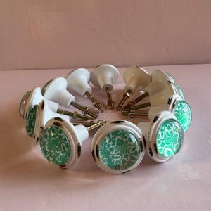 May include: A collection of white and green cabinet knobs arranged in a circle. The knobs have a white base with a distressed, brown rim and a clear, domed center featuring a green floral pattern. The hardware is visible.