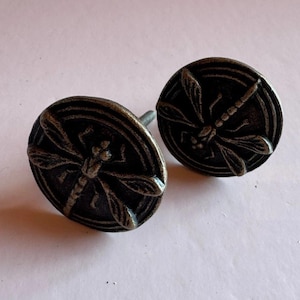 May include: Two antique bronze-colored cabinet knobs. Each knob features a detailed dragonfly design within a circular frame. The knobs have a distressed finish, adding to their vintage appeal. The hardware is suitable for drawers or cabinets.