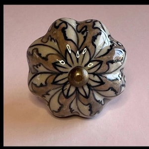 May include: A decorative ceramic cabinet knob with a floral design. The knob features a cream, brown, and black color scheme with a brass-colored center. The knob has a rounded, flower-like shape, perfect for adding a touch of elegance to furniture.