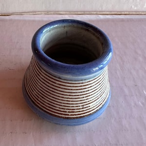 May include: A ceramic vase with a blue rim and base, featuring horizontal brown and white stripes. The vase has a flared shape, wider at the top and narrowing towards the base. The interior is a dark, neutral color.