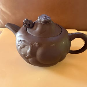 May include: A brown ceramic teapot with a rounded body, a curved spout, and a loop handle. The lid features a floral design and a small dragon-like figure. The teapot has an embossed floral pattern on its side.