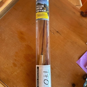 May include: A fly fishing rod in a clear plastic tube. The rod is a dark reddish-brown colour with a cork handle. The tube has a label that reads "YOUR NEW OFFICE" and "TFO". The tube also has a "Lifetime Warranty" label.