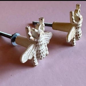 May include: Two off-white bee-shaped cabinet knobs with distressed detailing. Each knob features a threaded bolt for installation, a metal washer, and a cylindrical base. The knobs are set against a soft pink background.