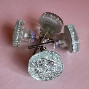 May include: Four clear glass cabinet knobs with various shapes, including round, square, and oval. Each knob has a decorative, textured design and is attached to a metal screw. The knobs are arranged on a pink surface.