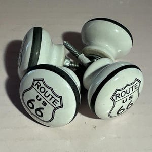 May include: Four white ceramic cabinet knobs with a Route 66 shield design in black and white. The knobs have a rounded shape with a black border and a metal screw for installation. These decorative knobs are ideal for adding a vintage touch to furniture.