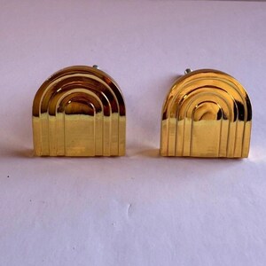 May include: Two gold-colored, art deco style earrings. Each earring has a half-circle shape with layered, arched ridges and vertical lines. The earrings are on a light purple background.