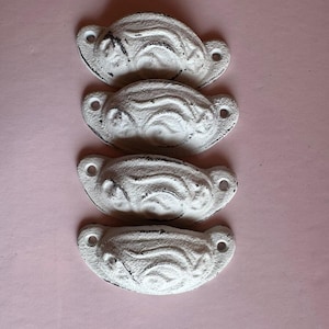 May include: Four white, distressed-finish drawer pulls with a curved, ridged design. Each pull has two holes for mounting. The pulls are arranged vertically against a pink background, suggesting a vintage or farmhouse aesthetic.