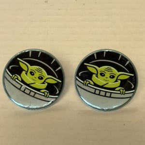 May include: Two round, silver-rimmed pins featuring a cartoon image of Grogu, also known as Baby Yoda, from the Star Wars franchise. The character is depicted in a pod against a black background.
