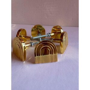May include: Set of gold-colored cabinet door pulls with a half-circle design. Each pull features a series of curved lines, creating an arched appearance. The pulls are attached to silver-colored screws and washers. The hardware is displayed on a light pink surface.
