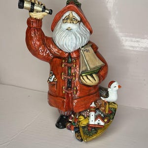 May include: A ceramic Santa Claus figurine in a red coat, holding a telescope and a sailboat. The figurine has a white beard and a nautical-themed shield with a lighthouse and a seagull.