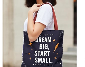 May include: Navy blue tote bag with the text "DREAM BIG, START SMALL." The bag features a starry design with an astronaut illustration and a lightning bolt. It has a red strap and is carried over the shoulder.