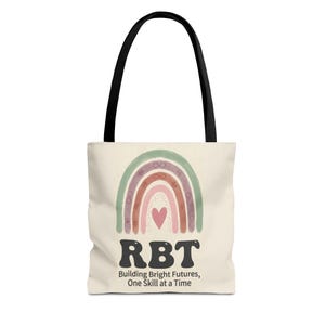 Puede incluir: Bolso tote color crema con asa negra. Presenta un diseño de arcoíris en tonos verdes, morados y rosas. Las letras "RBT" están en negrita negra, con el texto "Building Bright Futures, One Skill at a Time" debajo.