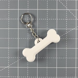 May include: A white bone-shaped keyring with a silver chain and ring. The bone is rounded and made of a solid, opaque material. The keyring is designed for pet owners.