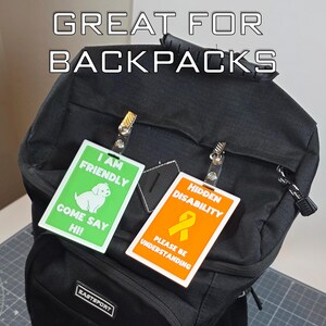 May include: Black backpack with two rectangular tags clipped to it. One tag is green with a white dog graphic and the text "I AM FRIENDLY COME SAY HI!". The other tag is orange with a yellow ribbon graphic and the text "HIDDEN DISABILITY PLEASE BE UNDERSTANDING".