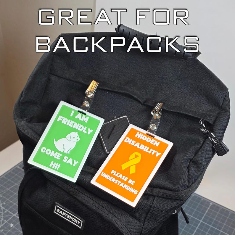 May include: Black backpack with two rectangular tags clipped to it. One tag is green with a white dog graphic and the text "I AM FRIENDLY COME SAY HI!". The other tag is orange with a yellow ribbon graphic and the text "HIDDEN DISABILITY PLEASE BE UNDERSTANDING".