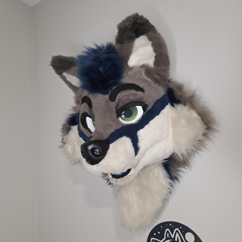 May include: A plush wolf head with a gray and white fur design, accented with dark blue details around the eyes and on the ears. The wolf has green eyes and a black nose. The head is mounted on a wall.