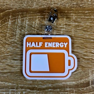 May include: An orange and white badge reel with the words "HALF ENERGY" and a battery icon showing half full. The badge reel has a metal clip and a clear plastic strap.