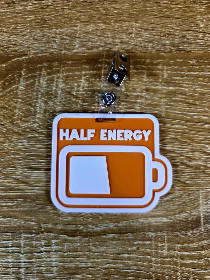 May include: An orange and white badge reel with the words "HALF ENERGY" and a battery icon showing half full. The badge reel has a metal clip and a clear plastic strap.