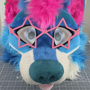 May include: A colorful furry wolf head with pink star-shaped glasses. The head is primarily blue with pink accents and a teal nose. The eyes are large and light-colored, and the mouth is slightly open, revealing a pink tongue.