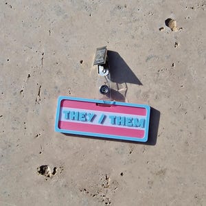 Badge showing they/them in trans colours on a tile background.
