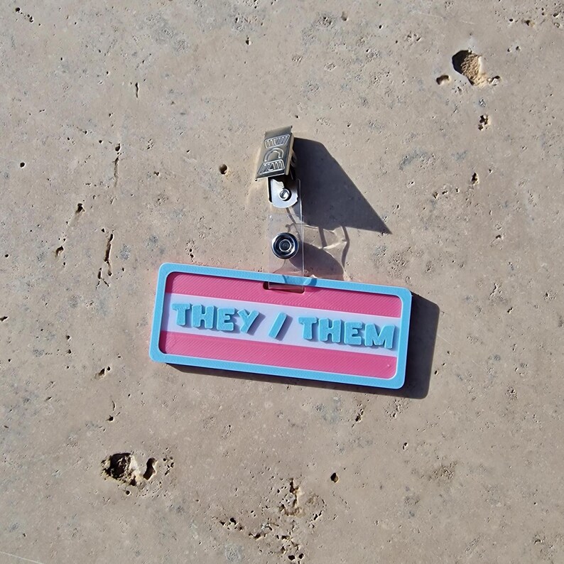 Badge showing they/them in trans colours on a tile background.