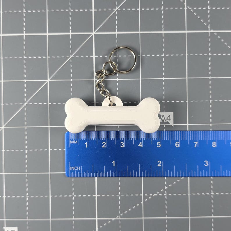 May include: A white, bone-shaped keyring, approximately 7.6 cm long, with a silver keyring and chain. The keyring is made of a solid, smooth material and is designed to resemble a dog bone. The background is a gray cutting mat with a blue ruler.