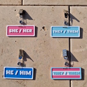 Pronoun badges on display on a tile background, badges displaying she/her, they/them, he/him and a they/them in the trans colours.