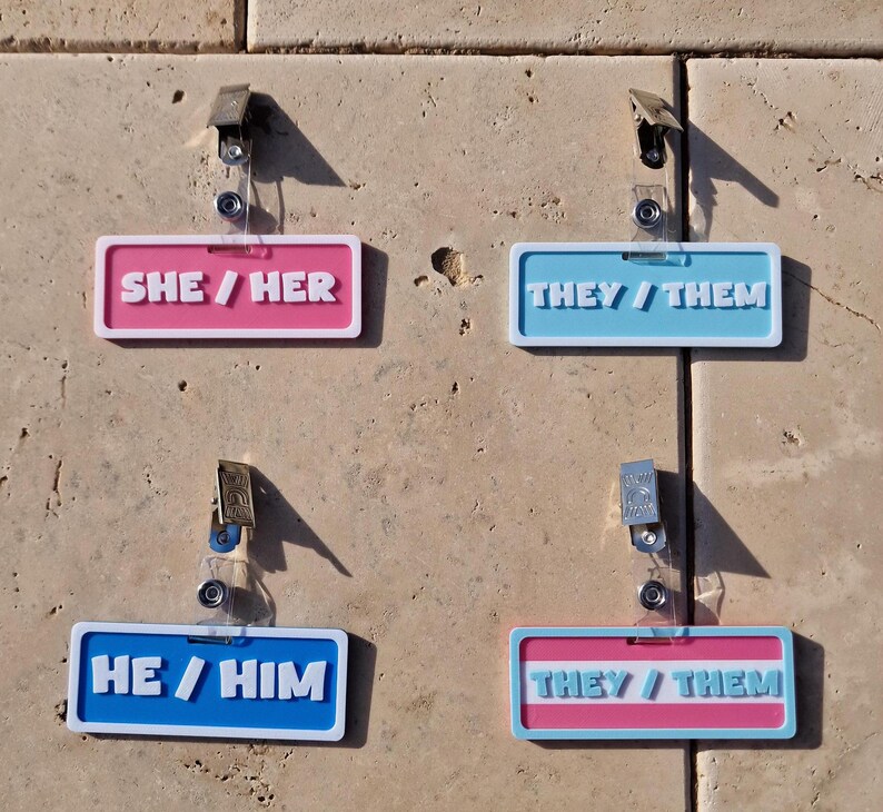 Pronoun badges on display on a tile background, badges displaying she/her, they/them, he/him and a they/them in the trans colours.