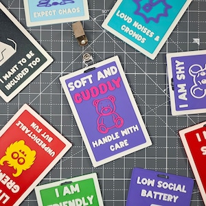 May include: A collection of colorful, rectangular badges with various phrases and graphics. One badge reads "SOFT AND CUDDLY" with a teddy bear illustration. Other badges include "I AM SHY," "LOW SOCIAL BATTERY," and "EXPECT CHAOS."