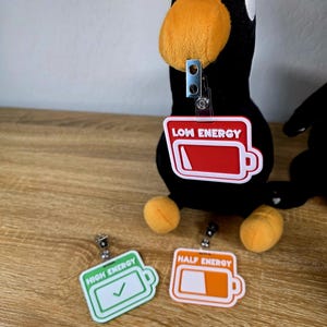 May include: Black plush penguin with orange beak and feet, and a badge reel. The badge reels have the text "LOW ENERGY", "HALF ENERGY", and "HIGH ENERGY" with battery graphics. The "HIGH ENERGY" badge has a checkmark.