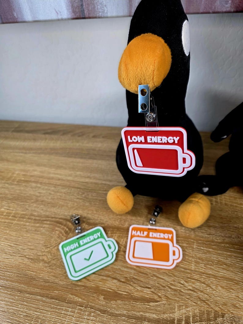 May include: Black plush penguin with orange beak and feet, and a badge reel. The badge reels have the text "LOW ENERGY", "HALF ENERGY", and "HIGH ENERGY" with battery graphics. The "HIGH ENERGY" badge has a checkmark.