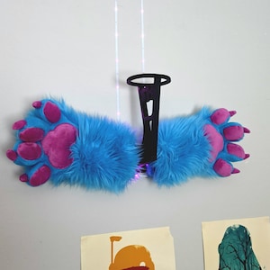 May include: A pair of plush blue and pink furry paw slippers. The slippers have large pink paw pads and claws. The slippers are hanging on a wall with a black decorative piece and string lights.