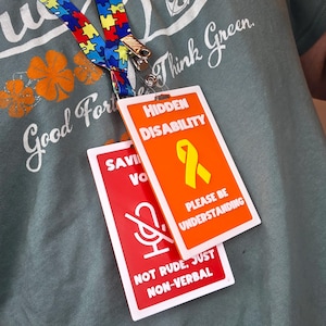 May include: A lanyard with a colorful puzzle piece pattern and a metal clip holding two rectangular badges. One badge is orange with the words "HIDDEN DISABILITY" and a yellow ribbon. The other badge is red with a microphone symbol and the words "NOT RUDE, JUST NON-VERBAL."