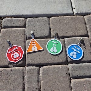 Op de afbeelding: Vier kleurrijke badges met knuffelthema met metalen clips. De badges hebben de vorm van een stopbord, een driehoek en twee cirkels. Het stopbord zegt "NO HUGS", de driehoek zegt "ASK FOR HUGS", en de cirkels zeggen "HUGS OK" en "HUGS PLEASE!"
