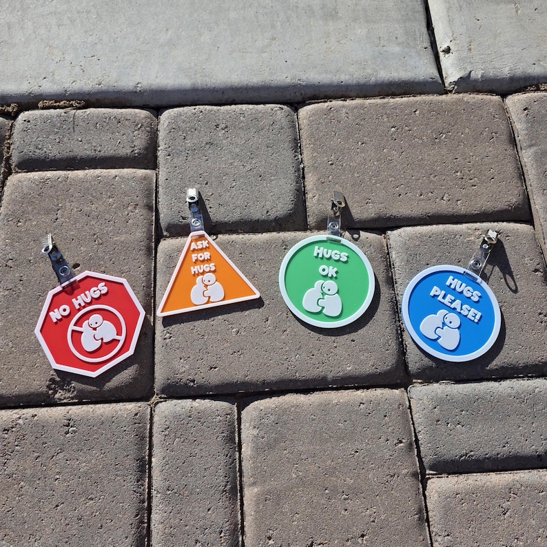 Op de afbeelding: Vier kleurrijke badges met knuffelthema met metalen clips. De badges hebben de vorm van een stopbord, een driehoek en twee cirkels. Het stopbord zegt "NO HUGS", de driehoek zegt "ASK FOR HUGS", en de cirkels zeggen "HUGS OK" en "HUGS PLEASE!"