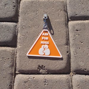 Op de afbeelding: Een oranje en witte driehoekige badge met de tekst "ASK FOR HUGS" en een afbeelding van twee figuren die elkaar omarmen. De badge is bevestigd aan een metalen clip. De achtergrond is een gestructureerd, grijs oppervlak.