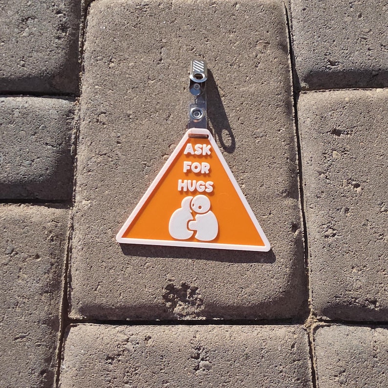 Op de afbeelding: Een oranje en witte driehoekige badge met de tekst "ASK FOR HUGS" en een afbeelding van twee figuren die elkaar omarmen. De badge is bevestigd aan een metalen clip. De achtergrond is een gestructureerd, grijs oppervlak.