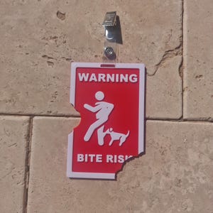 May include: A red and white warning sign with the text "WARNING BITE RISK" and a graphic of a person and dog. The sign is attached to a wall with a clear plastic clip.