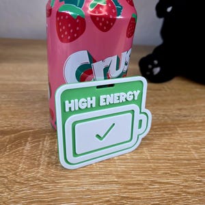 May include: A pink strawberry Crush soda can with a green and white logo, next to a green and white rectangular object with the words "HIGH ENERGY" and a battery icon with a checkmark. The can and object are on a wooden surface.
