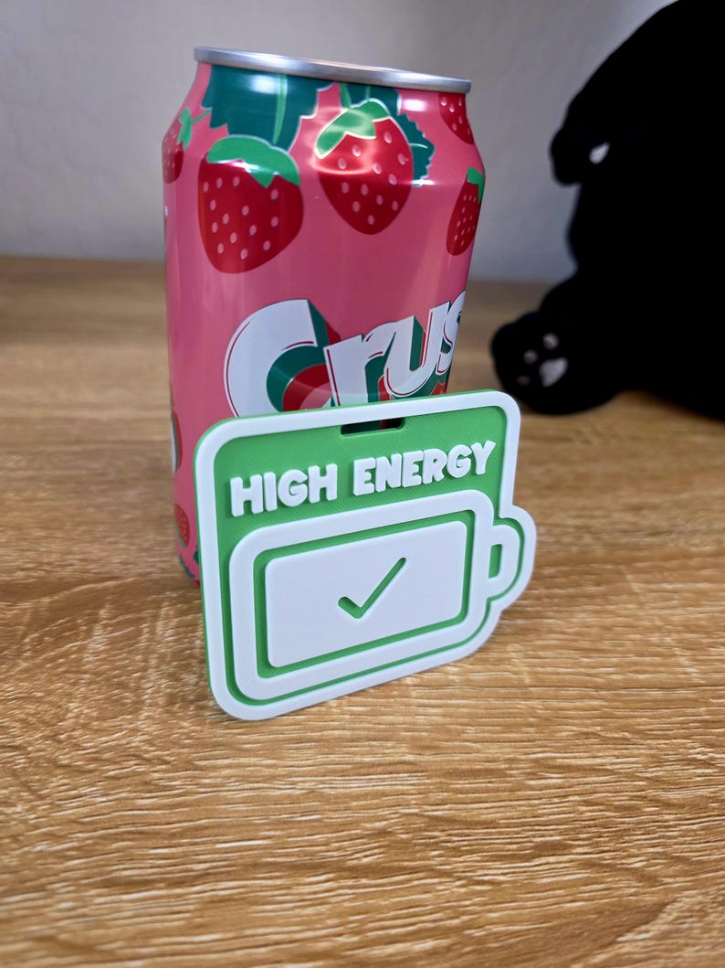 May include: A pink strawberry Crush soda can with a green and white logo, next to a green and white rectangular object with the words "HIGH ENERGY" and a battery icon with a checkmark. The can and object are on a wooden surface.