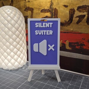 K&ouml;nnte beinhalten: Ein kleiner wei&szlig;er Staffelei-St&auml;nder h&auml;lt ein Schild mit den Worten "SILENT SUITER" &uuml;ber einem Lautsprechersymbol mit einem "X" hindurch. Das Schild ist blau mit wei&szlig;em Text und Grafiken. Links vom Schild befindet sich eine wei&szlig;e, strukturierte Lampe.
