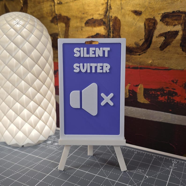 K&ouml;nnte beinhalten: Ein kleiner wei&szlig;er Staffelei-St&auml;nder h&auml;lt ein Schild mit den Worten "SILENT SUITER" &uuml;ber einem Lautsprechersymbol mit einem "X" hindurch. Das Schild ist blau mit wei&szlig;em Text und Grafiken. Links vom Schild befindet sich eine wei&szlig;e, strukturierte Lampe.