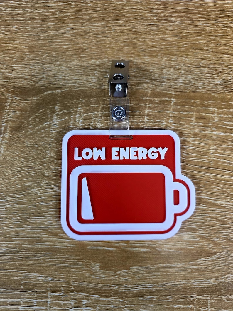 May include: A red and white name tag with the words "LOW ENERGY" above a battery icon. The battery icon is red with a white outline, indicating low power. A metal clip is attached to the top.