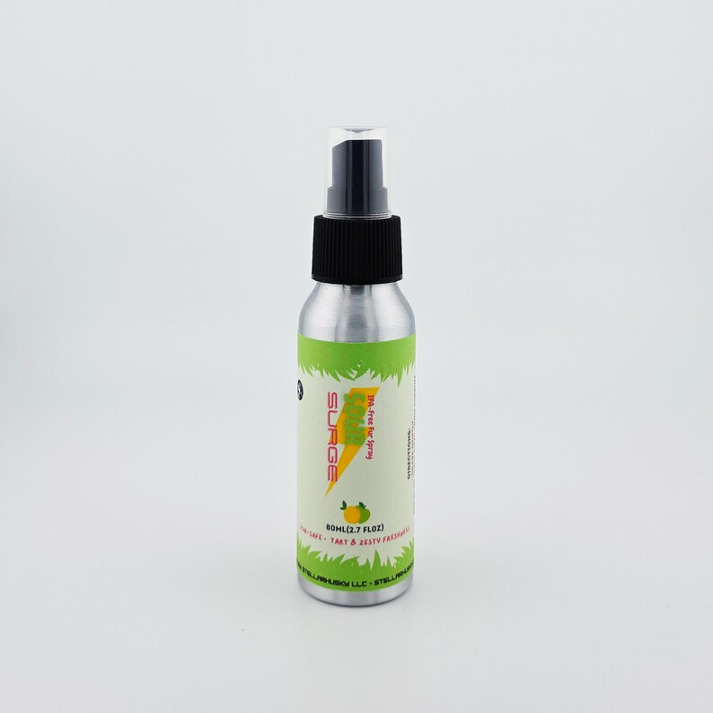 May include: A silver spray bottle with a black nozzle and cap. The label features the word "SURGE" in yellow, a lightning bolt graphic, and the text "The Fine Air Spray". The bottle contains 80ml (2.7 fl oz) of product.