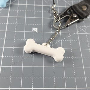 May include: A white bone-shaped dog tag suspended from a silver chain and a black strap with a metal clasp. The tag is made of a solid, smooth material. The background is a grey cutting mat with grid lines.