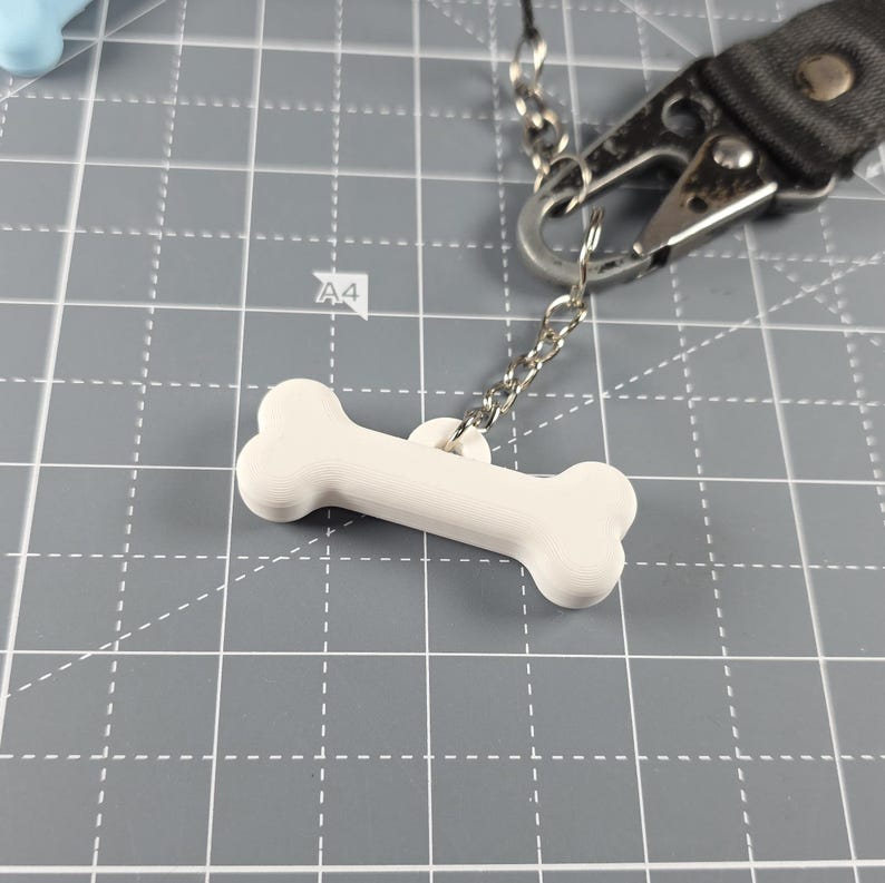 May include: A white bone-shaped dog tag suspended from a silver chain and a black strap with a metal clasp. The tag is made of a solid, smooth material. The background is a grey cutting mat with grid lines.