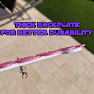 May include: A white and pink keychain with the text "THICK BACKPLATE FOR BETTER DURABILITY". The keychain has several pink circular accents and a silver clasp. The design is set against a blurred outdoor background.
